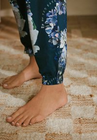 Jogginghosen mit Blumenmuster aus dunkelgrünem Stoff, ausgestattet mit einem elastischen Bündchen am Knöchel. Der Fuß ist barfuß auf einem strukturierten gewebten Teppich mit beigen und weißen Quadraten.