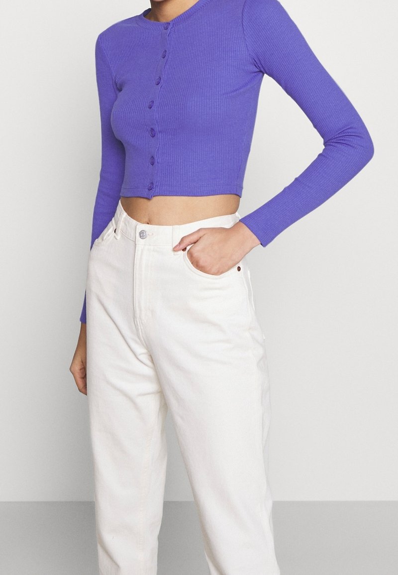 Långärmad lila ribbad croppad topp med rund halsringning och knäppning framtill, matchad med högmidjade vita jeans.