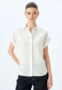 Camicia bianca a maniche corte con bottoni, finitura tessurizzata e colletto a punta, abbinata a jeans scuri. Design pulito e minimale.