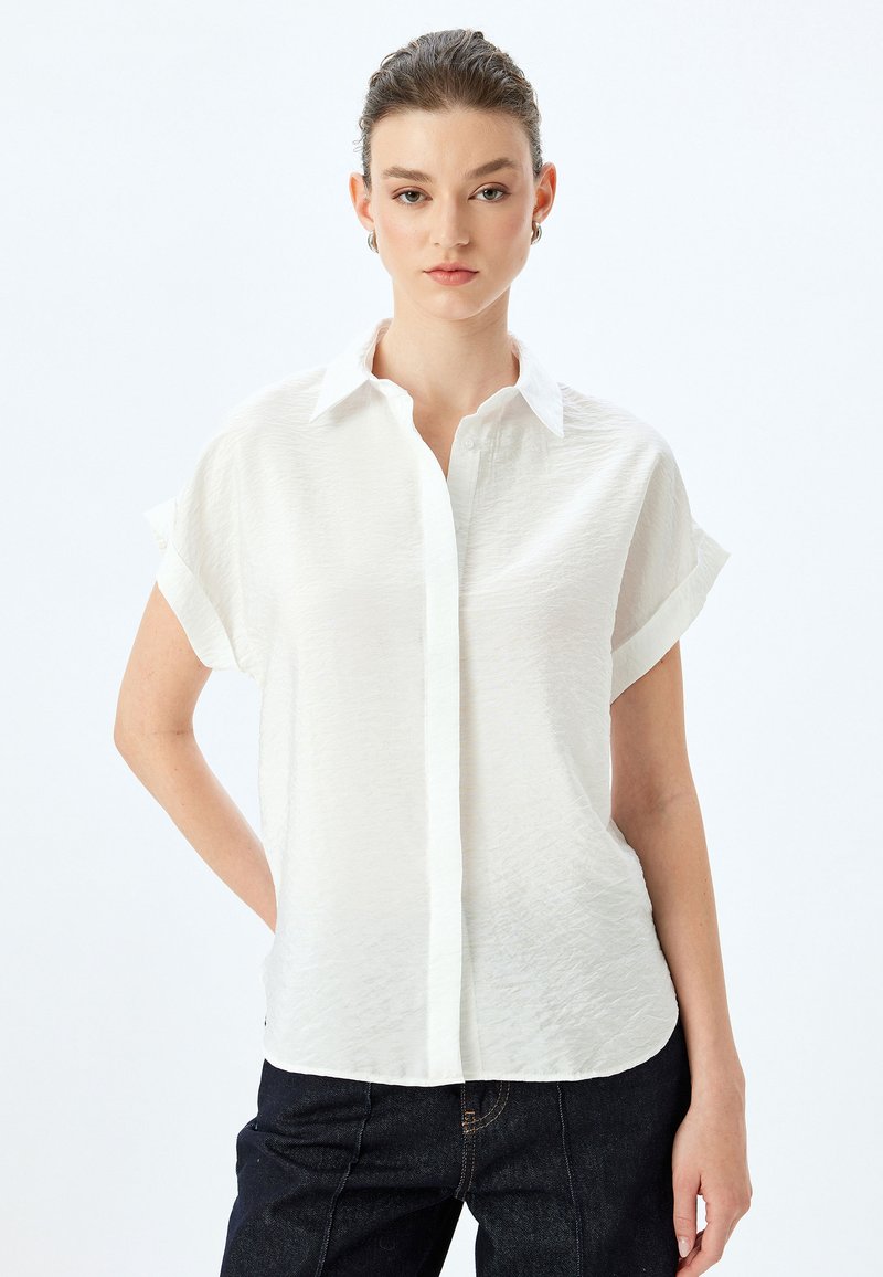 Camisa de botón de manga corta blanca con un acabado texturizado y cuello puntiagudo, combinada con jeans oscuros. Diseño limpio y minimalista.
