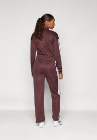 Burgundy idrottsoverall med vita sidostreck, tillverkad av mjukt tyg. Har en figurnära jacka och löst sittande byxor, bärs med vita sneakers.