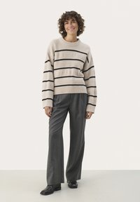 Pull en beige à rayures noires, avec un col et des poignets côtelés, associé à un pantalon large gris et des chaussures noires. Vue de face.