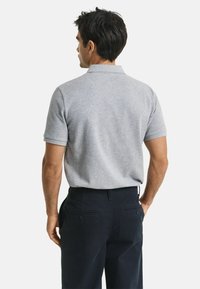 Mann in einem hellgrauen Polohemd mit kurzen Ärmeln und dunkeln Marineblauen Hosen, der mit dem Rücken zur Kamera vor einem schlichten Hintergrund steht.