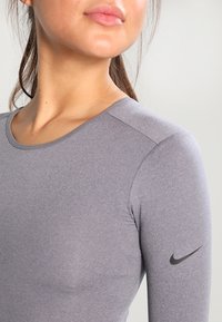 Grå langærmet atletisk top med rund halsudskæring og tætsiddende design. Har et sort Nike-logo på venstre ærme. Glat tekstur.