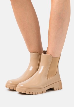 Vadderade, glänsande beige gummistövlar som går upp till anklarna med kraftiga sulor, bärs på bara ben mot en enkel vit bakgrund.