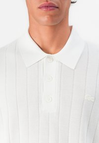 Close-up of a person wearing a white ribbed knit polo shirt with three buttons and a small embroidered crocodile logo on the chest.