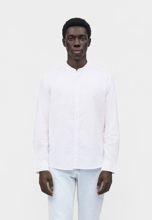 Jeune homme à la peau foncée et aux cheveux courts et bouclés, portant une chemise blanche à col mandarin et un jean bleu clair, debout devant un fond uni.