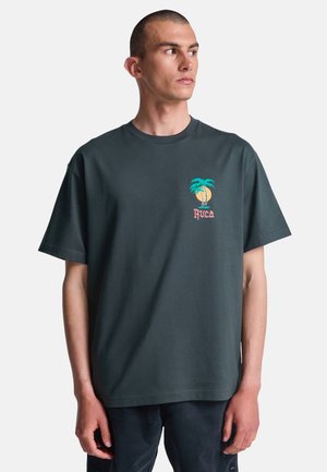 Mężczyzna w ciemnoszarej koszulce z małą kolorową grafiką palmy i zachodu słońca oraz napisem "RVCA" na lewej piersi, patrzący w prawo.