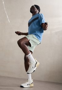 Zila sporta tees ar melniem akcentiem, gaiši zaļi šorti, baltas teksturētas zeķes un baltas/krēmkrāsas Nike apavi ar melniem elementiem. Rokā ir lecamauklas.