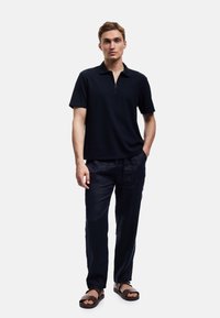 Granatowa koszulka polo z krótkim rękawem i zamkiem błyskawicznym przy kołnierzu, w zestawieniu z granatowymi lnianymi spodniami i brązowymi płaskimi sandałami. Gładka tekstura, luźny krój.