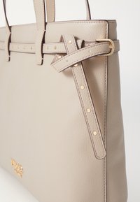 Sac cabas en cuir beige avec poignées doubles, détails à clous et accent boucle dorée. Présente une surface texturée et un design spacieux.