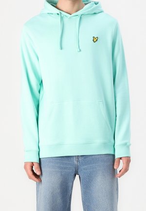 Mann trägt hellblauen Kapuzenpullover mit Fronttasche und gelbem Vogel-Logo auf der Brust, kombiniert mit blauen Jeans.