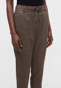 Person trägt braune Strickhose mit Vordertaschen und Kordelzugbund, kombiniert mit einem schwarzen Oberteil vor schlichtem hellen Hintergrund.