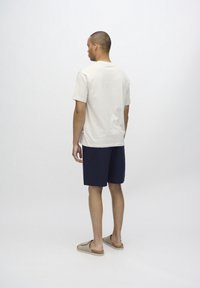 T-shirt en coton beige clair, short bleu marine avec un motif texturé, et chaussures slip-on beiges. Jambes nues et fond neutre.