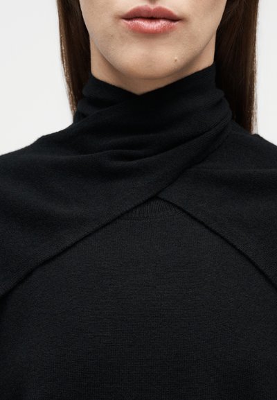 Κοντινό πλάνο ενός ατόμου που φορά ένα μαύρο πλεκτό πουλόβερ με ψηλό, διασταυρωμένο γιακά που καλύπτει τον λαιμό.