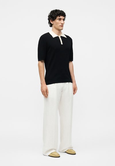 Homme debout, portant un polo noir à manches courtes avec un col blanc, un pantalon blanc ample et des mocassins beige sur un fond clair uni.