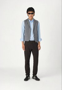 Siva prsluk preko svijetloplave košulje, crne hlače i crne tenisice s bijelim prugama. Model nosi tamne sunčane naočale.