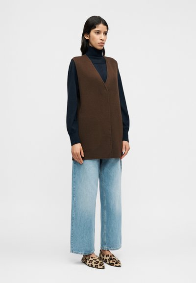 Καφέ πλεκτό γιλέκο χωρίς μανίκια με V-λαιμόκοψη, φορεμένο πάνω από ένα ναυτικό ζιβάγκο, συνδυασμένο με ανοιχτό μπλε τζιν με φαρδιά πόδια και φλατ παπούτσια με λεοπάρ σχέδιο.