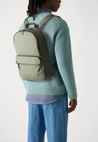 Mochila verde oliva hecha de material suave, con un bolsillo frontal con cremallera, correas acolchadas y un diseño minimalista. Llevada sobre un suéter azul claro.