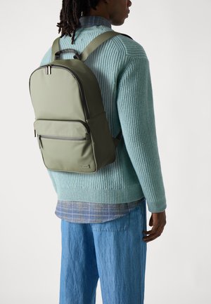 Mochila verde oliva hecha de material suave, con un bolsillo frontal con cremallera, correas acolchadas y un diseño minimalista. Llevada sobre un suéter azul claro.