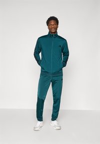 Turquoise sportoutfit gemaakt van gladde stof, bestaande uit een ritsjack met zwarte strepen en logo, gecombineerd met bijpassende tapered broek en witte sneakers.