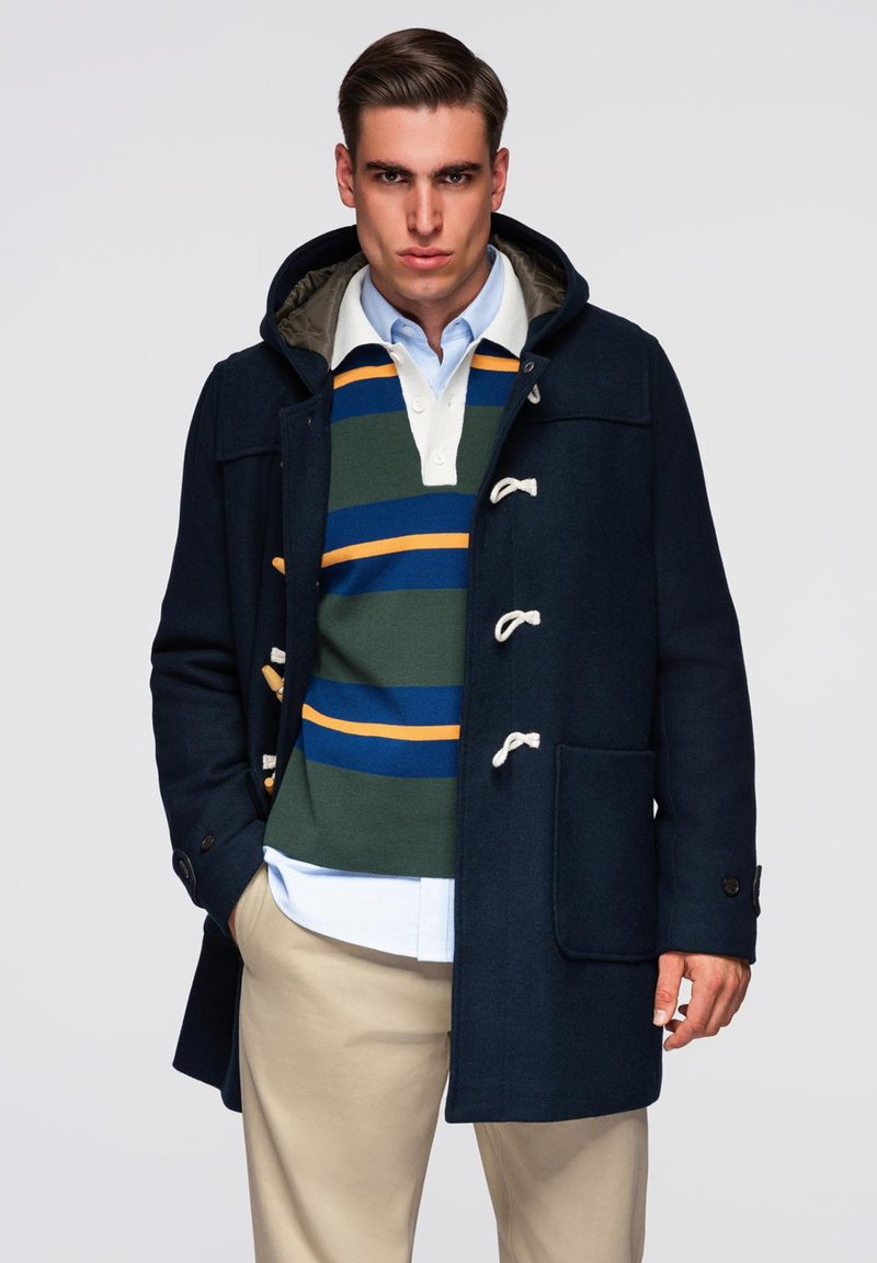 Mørkeblå uld duffeljakke med toggle-lukninger, hætte og forlommer, lagdelt over en grøn og blå stribet sweater med en orange stribe.