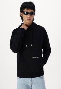 Hoodie noir avec une poche avant, un capuchon à cordon et un logo blanc. Porté par-dessus un jean bleu clair. Le modèle porte également des lunettes de soleil noires.