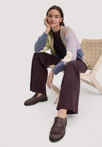 Bruine leren loafers met franjes, gecombineerd met donkerpaarse broek met wijde pijpen en een multi-kleurige gebreide cardigan. Zittend op een houten stoel.