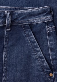 Primo piano di jeans in denim blu scuro che mostra la vita, l'asola per la cintura, la tasca anteriore, le cuciture e il dettaglio della rivettatura in metallo.