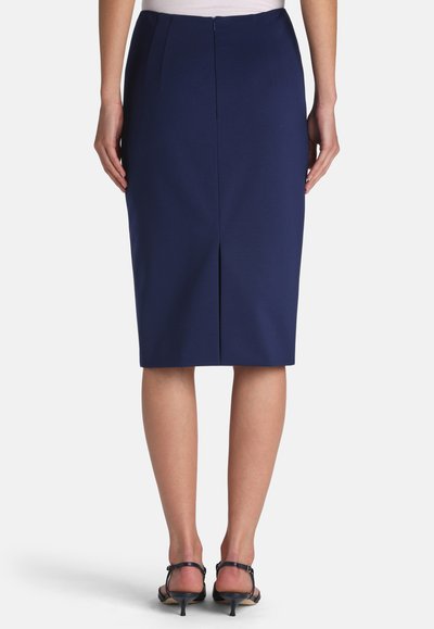Woman wearing a navy blue knee-length pencil skirt with back slit, paired with black low-heeled sandals, standing against a white background.