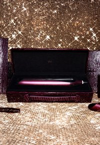 Ghd Haarglätter in einem dunkellila strukturierten Etui, ausgekleidet mit schwarzem Samt, auf einem glitzernden goldenen Hintergrund präsentiert.