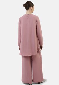 Tunika rosa e testurizzata con maniche lunghe e dettaglio a chiave sulla schiena; abbinata a pantaloni a gamba larga. Silhouette semplice e morbida.