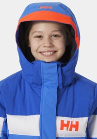 Modrá izolační bunda s jasně oranžovým kapucí. Má horizontální bílé pruhy a výrazné logo na hrudi. Hladká textura.