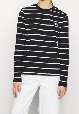 Persoon draagt een zwart shirt met lange mouwen met witte horizontale strepen en de tekst "OBEY" op de borst, gecombineerd met een witte broek.