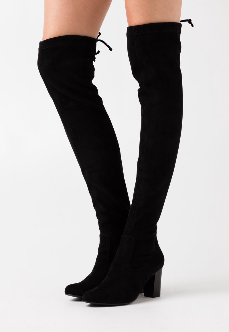 Bottes en daim noir au-dessus du genou avec talons blocs portées sur jambes nues sur fond blanc uni.
