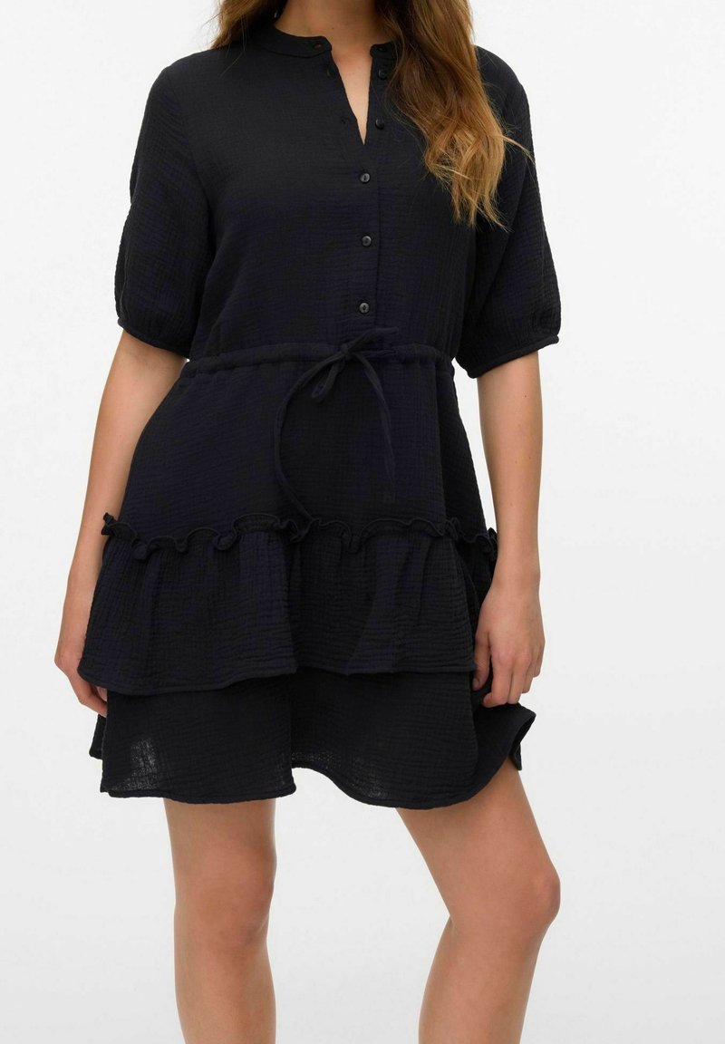Femme portant une robe noire texturée à manches courtes avec des boutons, une ceinture nouée à la taille et un ourlet à volants, debout devant un fond blanc uni.