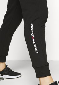 Mustat collegehousut, joissa on resorireunukset ja valkoinen kylkilogo, jossa lukee "TOMMY HILFIGER" punaisella ja sinisellä korostuksella. Pehmeää, joustavaa kangasta.