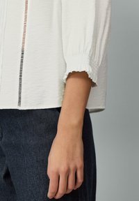 Blouse blanche texturée avec des manches évasées et froncées, dotée de découpes verticales. Associée à un jean en denim foncé.