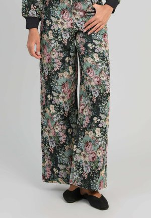 Pantalon à jambes larges avec fond sombre et motif floral multicolore, porté avec des chaussures noires sans lacets et une main posée sur la hanche.