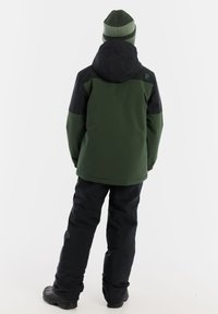 Grüne und schwarze isolierte Jacke mit Kapuze, getragen über schwarzen Schneehosen. Das Material scheint wasserdicht zu sein, mit sichtbaren Nähten und Logodetails.