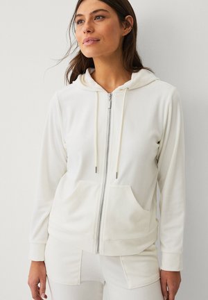 Frau trägt weißen Kapuzenpullover mit Reißverschluss, Vordertaschen und Kordelzug, steht vor einer schlichten hellen Wand und schaut leicht zur Seite.