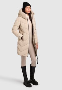 Beige pufferjakke med hætte, med et chevronmønster, sidelommer og sort lynlås. Bæres med ribbede beige leggings og sorte knælange støvler.