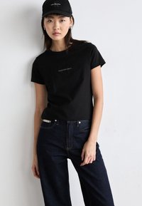 Juoda medvilnės marškinėliai su trumpomis rankovėmis, pasižymintys mažu Calvin Klein Jeans logotipu. Derinami su tamsiai mėlynomis džinsais ir juoda kepure.