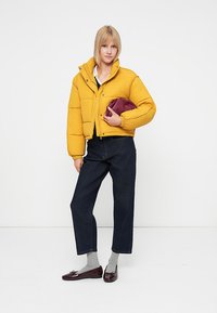 Gul cropped pufferjakke, lynlås med trykknapper, kombineret med mørke denimbukser og en bordeaux clutch. Grå sokker og mørke loafers.