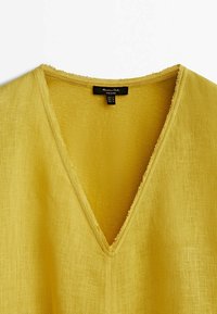 Haut en lin jaune avec un décolleté en V et des bords bruts, montrant le haut de la poitrine et l'étiquette intérieure avec les informations de taille.