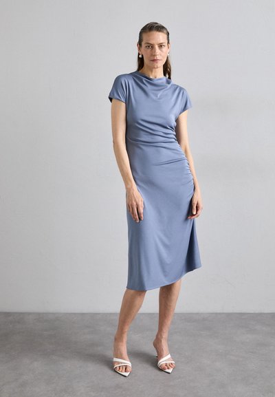 Blauwe stretch stof jurk met korte mouwen, een aansluitend ontwerp, gedrapeerde details en een zoom tot de knie. Gecombineerd met witte hakken met bandjes.