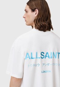 Hvid bomulds t-shirt med stort aqua tekst "ALLSAINTS" på ryggen, med ekstra japansk tekst og "LONDON" i en mindre skrifttype nedenfor.