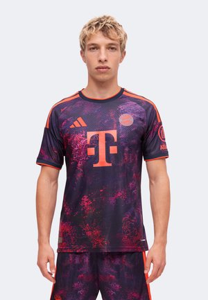 Junger Mann trägt ein dunkelviolettes und rotes, gemustertes Bayern München Fußballtrikot mit orangen Logos und passenden Shorts und steht vor einem schlichten Hintergrund.