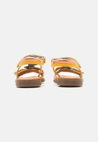 Zwei Sandalen im Rückansicht mit gelben, orangefarbenen und grünen Riemen, aus Leder und Stoff, mit braunen Gummisohlen und verstellbaren Schnallen.