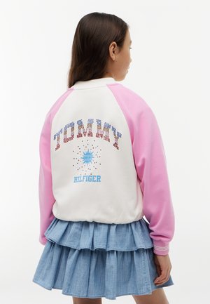 Mädchen trägt eine weiß-rosa Jacke mit dem "Tommy Hilfiger"-Logo und einen mehrlagigen blauen Jeansrock, mit dem Rücken zu einem schlichten Hintergrund.